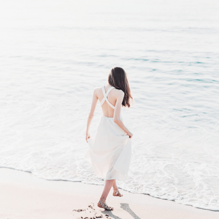 Woman standing barefoot in the sea, wearing a light dress, facing away. Symbol of calm, reflection, and inner peace.
