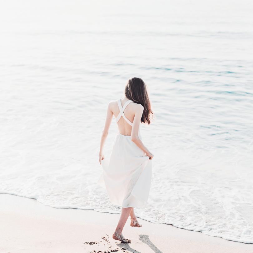 Woman standing barefoot in the sea, wearing a light dress, facing away. Symbol of calm, reflection, and inner peace.
